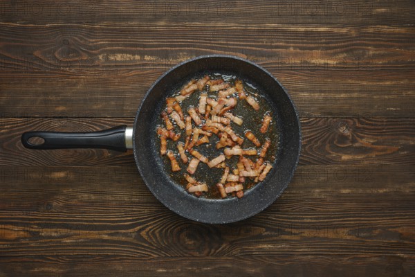 Sizzling strips of bacon fry in a black skillet, releasing savoury aroma and fat on rustic wooden