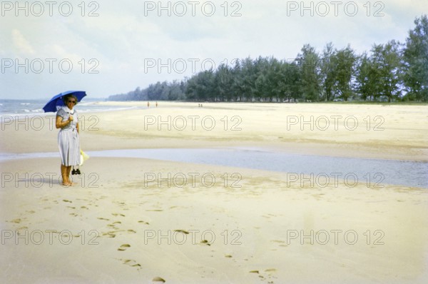 British woman holding umbrella standing on sandy beach south of estuary, Rompin, East coast,