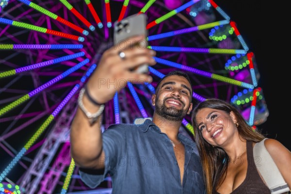 Young couple smiling and taking a selfie with smartphone in front of a colorful ferris wheel at an