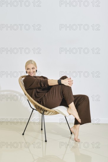 A barefoot woman dressed in brown lounge wear relaxes in a stylish chair, smiling and exuding a