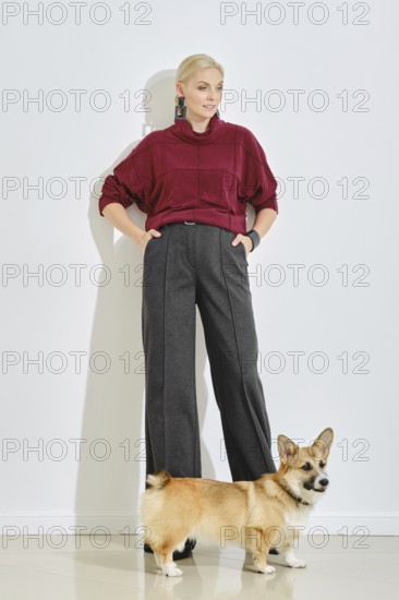 A woman dressed in a burgundy sweater and gray wide-leg pants stands confidently next to her corgi.