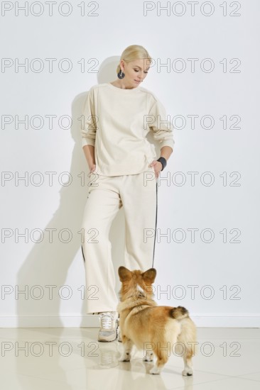 A woman dressed in light, casual attire stands against a plain wall while a corgi dog curiously
