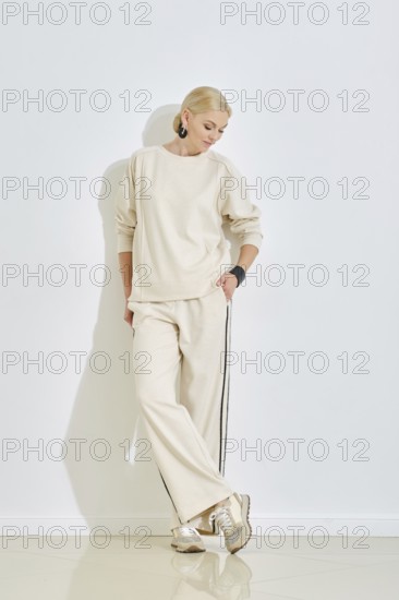 A woman dressed in a cozy beige outfit leans casually against a white wall, showcasing a relaxed