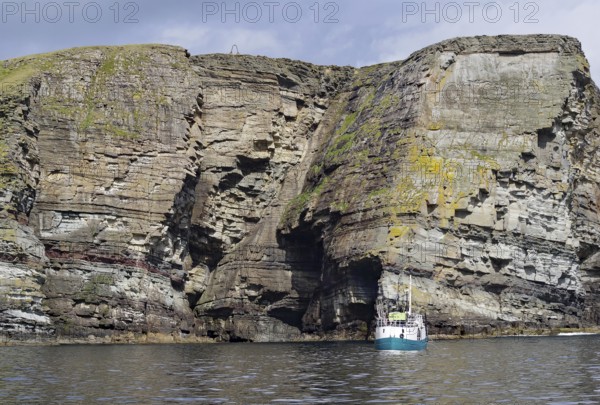 Boat in front of massive cliffs in the sea under a cloudy sky, Noss, Shetland Islands, Scotland,