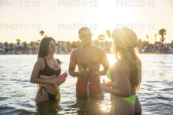 Three friends enjoying pineapple cocktails in the sea at sunset, celebrating summer vacation with