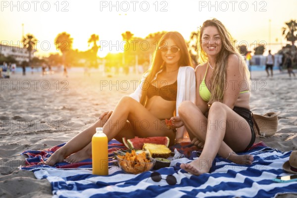 Two friends savoring a delightful picnic on the beach, surrounded by fresh fruit and juice, while