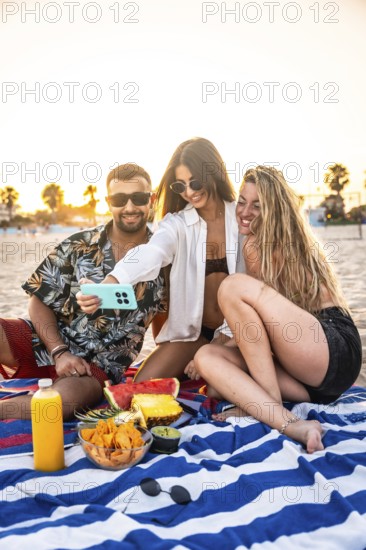 Three friends are taking a selfie with smartphone during a picnic at the beach, enjoying snacks and