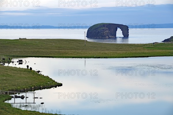 Calm lake with grassy shores and a rock formation on the horizon, dore holm Dore Holm, Hillswick,