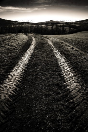 Tire tracks cut through a dark, rural field, leading toward rolling hills under a moody sky at dusk