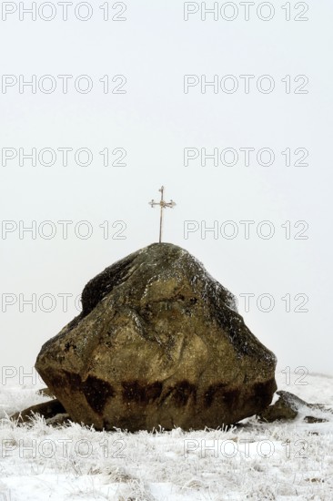A large rock topped with a cross stands starkly against the winter landscape of the Aubrac Plateau