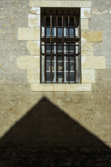 A barred window positioned on a rough stone wall casts a strong shadow below. Sunlight enhances the