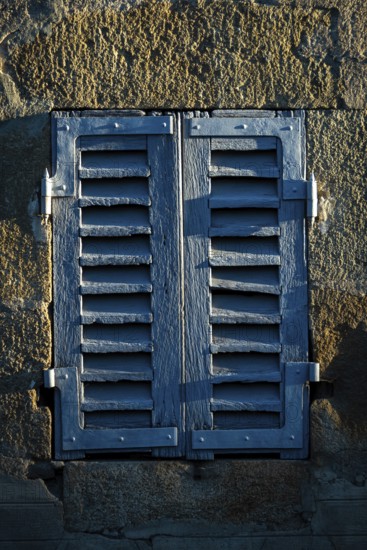Sunlight casts shadows on a pair of weathered blue shutters attached to an old stone wall. The