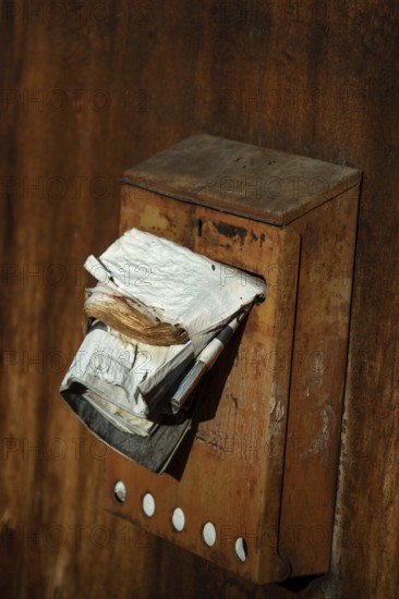 A rusty mailbox attached to a weathered wall is bursting with various papers and ads, indicating
