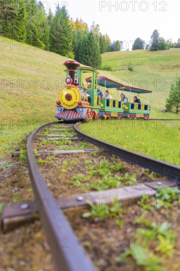 A colourful mini train moves through a green hilly landscape on narrow tracks, Retro Freizeitpark