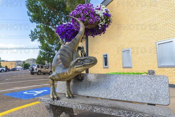 Dinosaur statue resting on a bench in front of a brick building, surrounded by flower baskets in