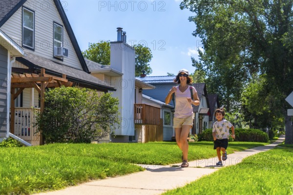 Mother and young son running joyfully on a sidewalk in a sunny, residential neighborhood,