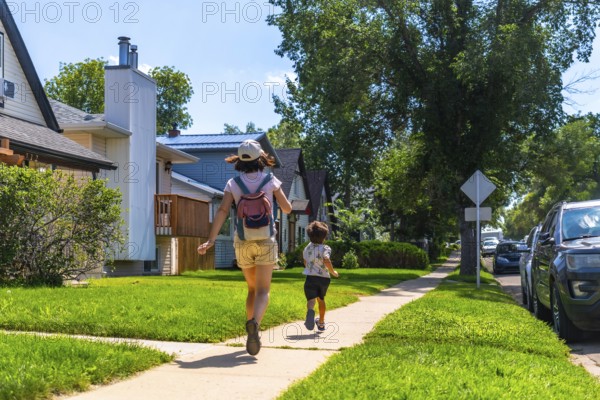Mother with backpack and her son are running down a sidewalk in a residential area of drumheller,