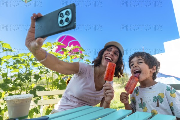 Mother and her son are sitting at a turquoise picnic table, holding popsicles and taking a selfie