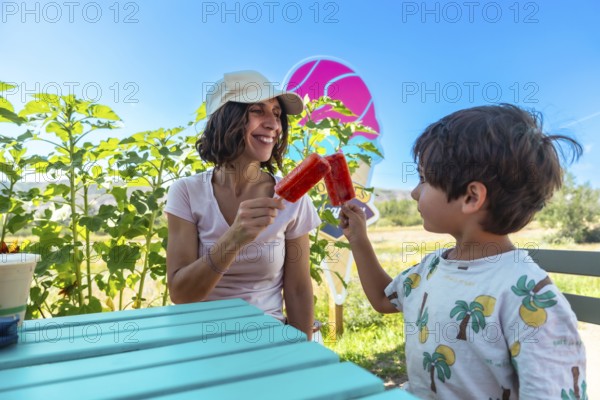 Mother and son are enjoying colorful popsicles at a picnic table on a sunny summer day in