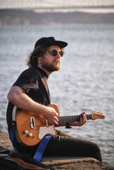 Hipster street musician in black playing electric guitar in the street sitting on pier embankment