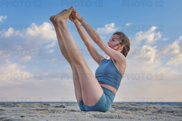 A woman in Ubhaya Padangusthasana, or Double Toe Hold, balancing gracefully on the sandy beach. The