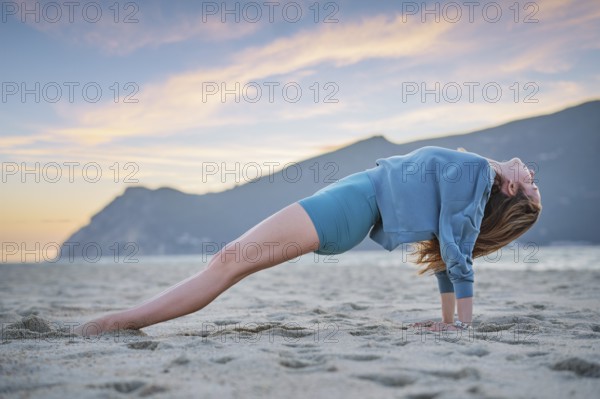 A woman practices Purvottanasana, or upward plank pose, on the beach during sunset. The golden sky