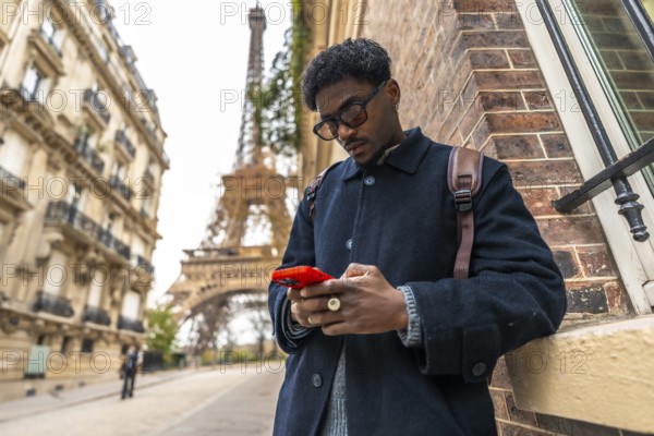 Young black man in paris leaning against a building, focusing on his smartphone, representing