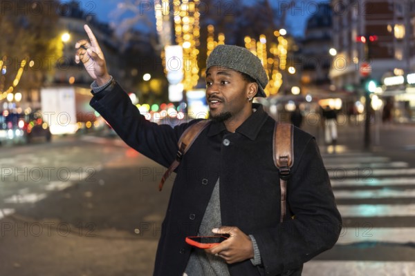Young man in a winter coat stands on a busy city street at night, holding a smartphone and raising