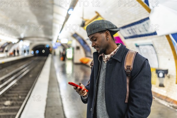 Young black man in a flat cap and backpack checks his smartphone while waiting on a busy urban