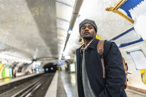Young black man in a beret with a backpack stands on a tiled paris metro platform, looking up while