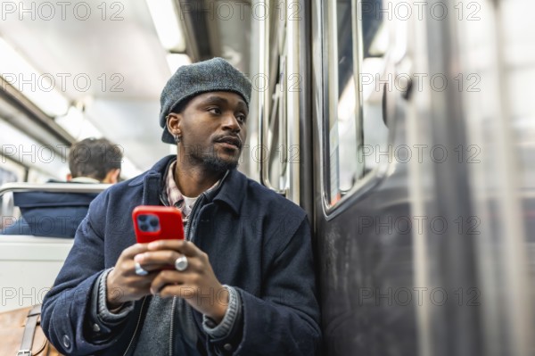 Young black man seated on subway, dressed for winter, holding smartphone and gazing out the window