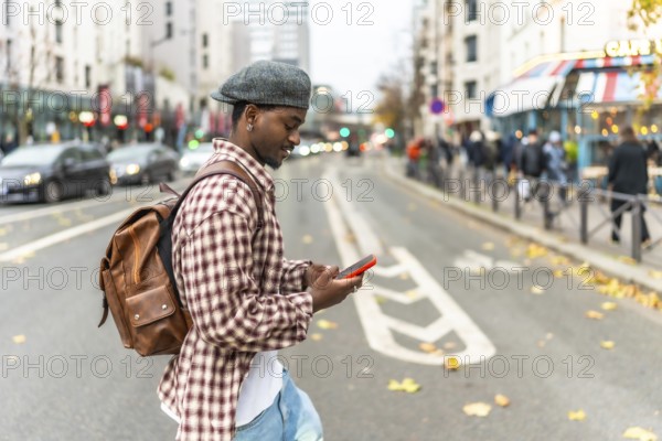 Young man wearing a beret and backpack walking across a city street, looking down at his smartphone