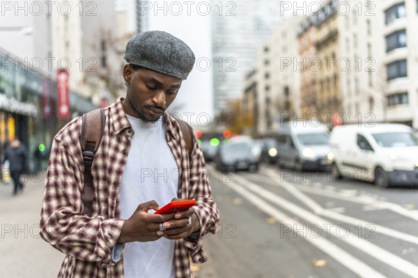 Young black man standing on an urban street, wearing a plaid shirt and a cap, engrossed in using