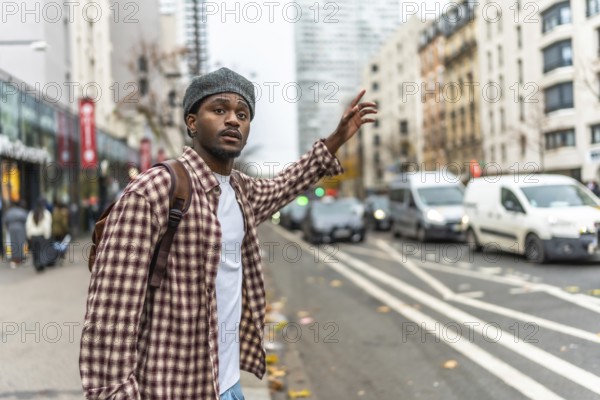 Young black man wearing a beret and a plaid shirt standing on an urban sidewalk, raising his hand