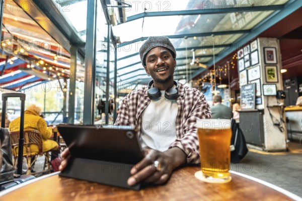 Young black man in a plaid shirt and hat, smiling at camera while enjoying a beer and using a