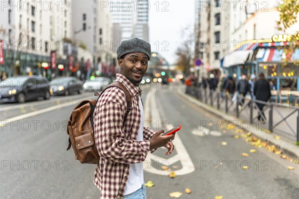 Young black man in a checkered shirt and backpack smiles at the camera while checking his phone on