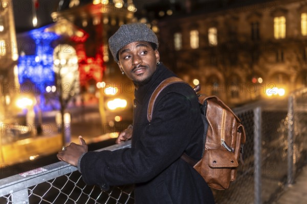 Young black man wearing a coat and a beret, carrying a backpack, standing on a bridge overlooking