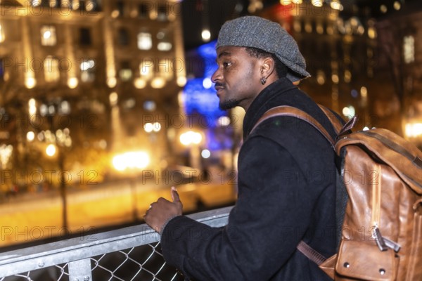 Young black man wearing a cap and coat with a brown backpack, standing on a bridge, thoughtfully