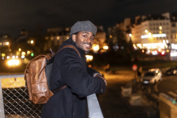 Cheerful male tourist wearing a classic cap and brown backpack, confidently posing on a bridge rail