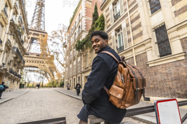 Happy black man tourist with a backpack sitting on a railing on a charming parisian street, smiling