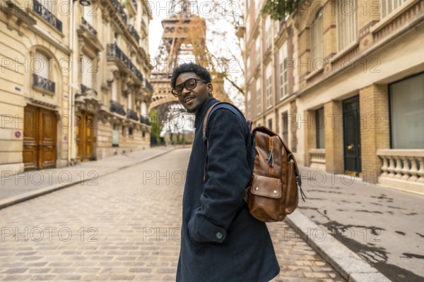 Young african man with glasses and backpack smiles confidently while walking a cobblestone paris