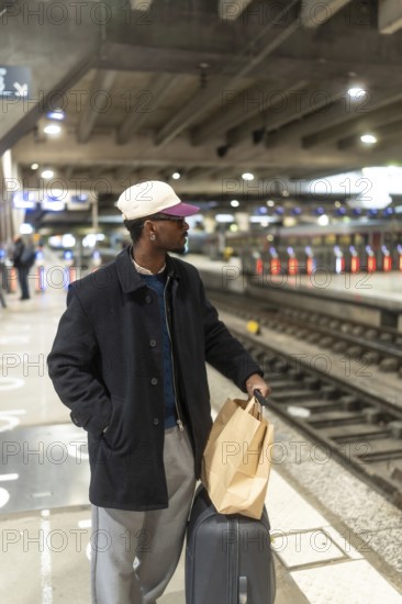 Young man with a baseball cap and glasses standing on a train station platform, holding a shopping