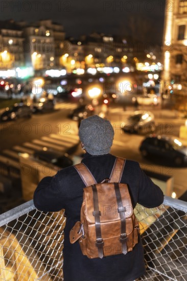 Person with a stylish leather backpack and knit cap viewing the illuminated cityscape of paris,