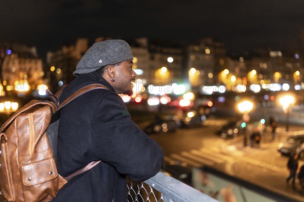 Young black man smiling and enjoying a stunning view of paris street lights at night, wearing a cap
