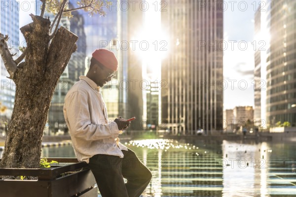 Young black man in casual attire concentrating on his smartphone while leaning by a tree, with