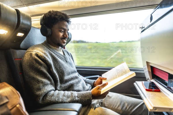 Young man wearing headphones and reading a book while traveling comfortably by high speed train,