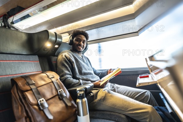 Happy black man wearing headphones and reading a book while traveling by train, sitting comfortably