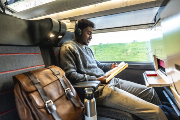Young black man traveling on a modern train, wearing headphones and reading a book, enjoying a