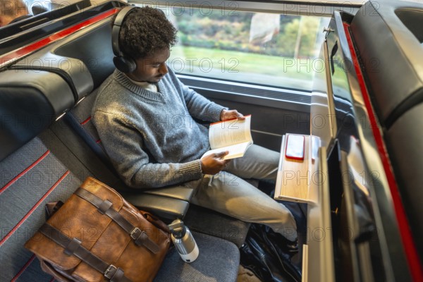 Young professional in headphones sits by a train window reading a book during a high speed