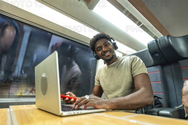 Young black man with headphones smiling while working on a laptop and using a smartphone during a